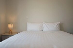 Una cama bien hecha con sábanas blancas, una lámpara de noche y una pared lisa.