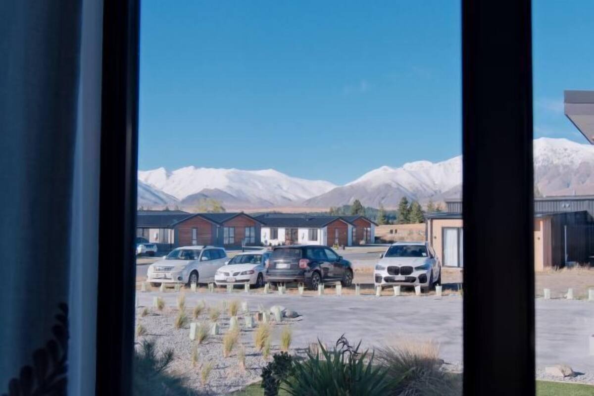 Blick auf einen Parkplatz mit mehreren Autos, ein Gebäude mit flachem Dach und verschneite Berge im Hintergrund.