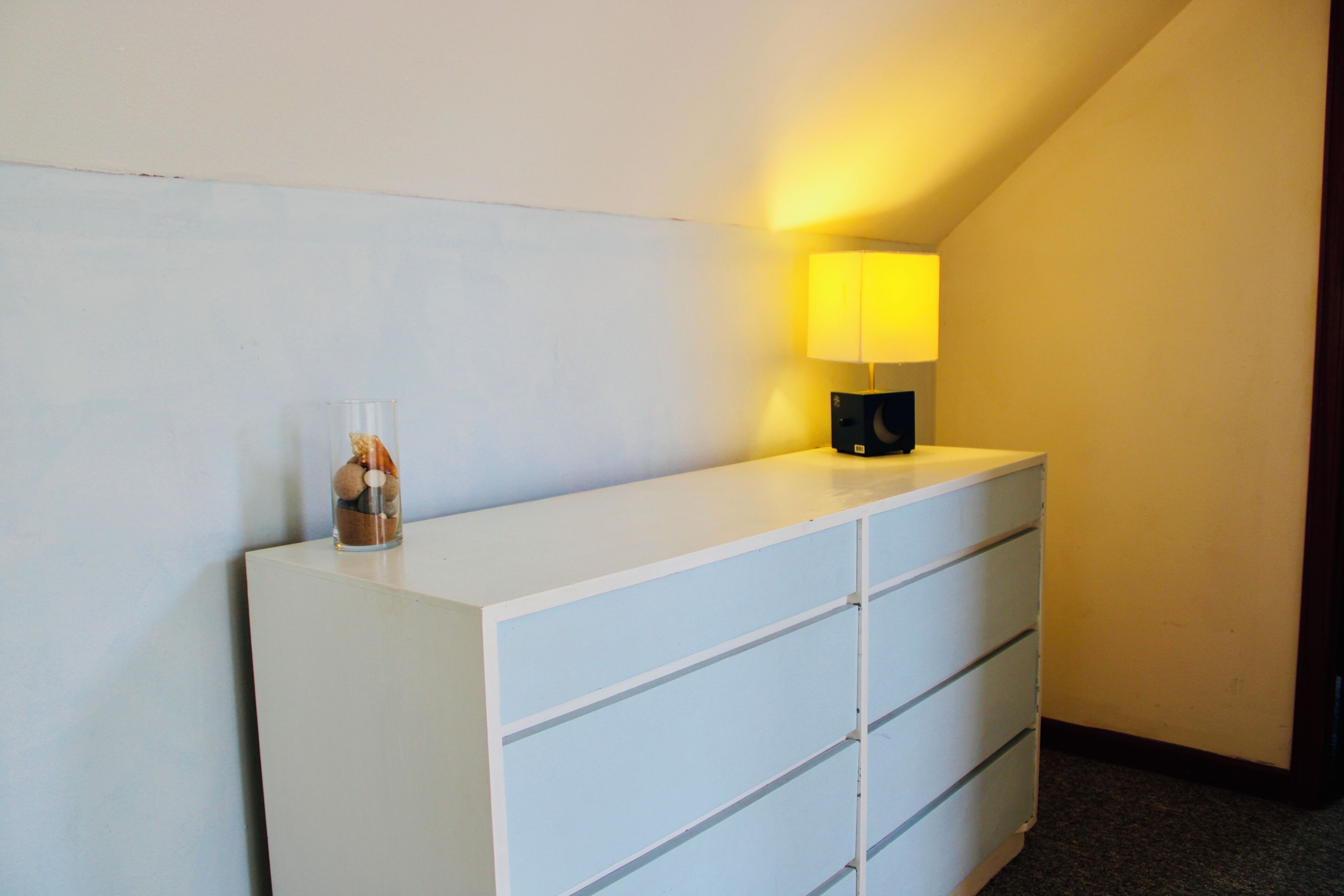 A white dresser with drawers and a glass container on top. A lamp with a yellow shade is on the dresser.