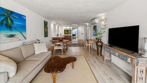 A spacious living room with a sofa, coffee table, and TV stand. There is a dining area with a table and chairs, and a kitchen in the background.