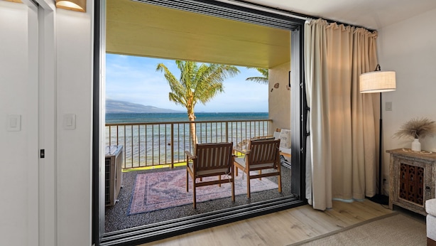A balcony with a view of the sea, two wooden chairs, a small table, a lamp, and a decorative cabinet.