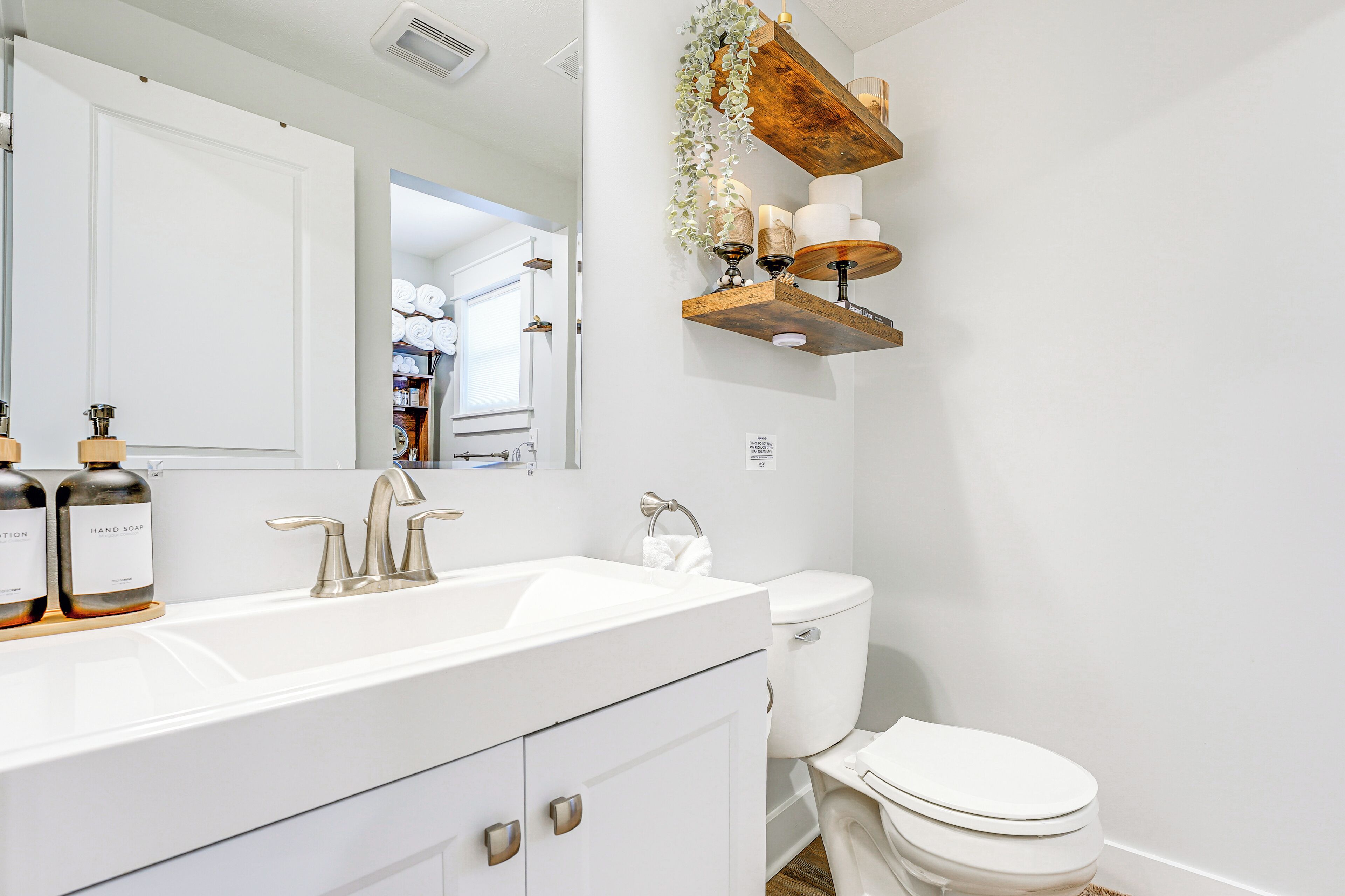 A modern bathroom with a white sink, a toilet, and a wooden shelf above the toilet holding toiletries.
