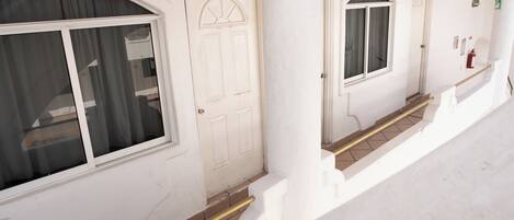 Un edificio blanco con tres puertas, cada una con un ventanal encima. Hay escalones que llevan a las puertas, y un extintor está montado en la pared cerca de la puerta derecha.