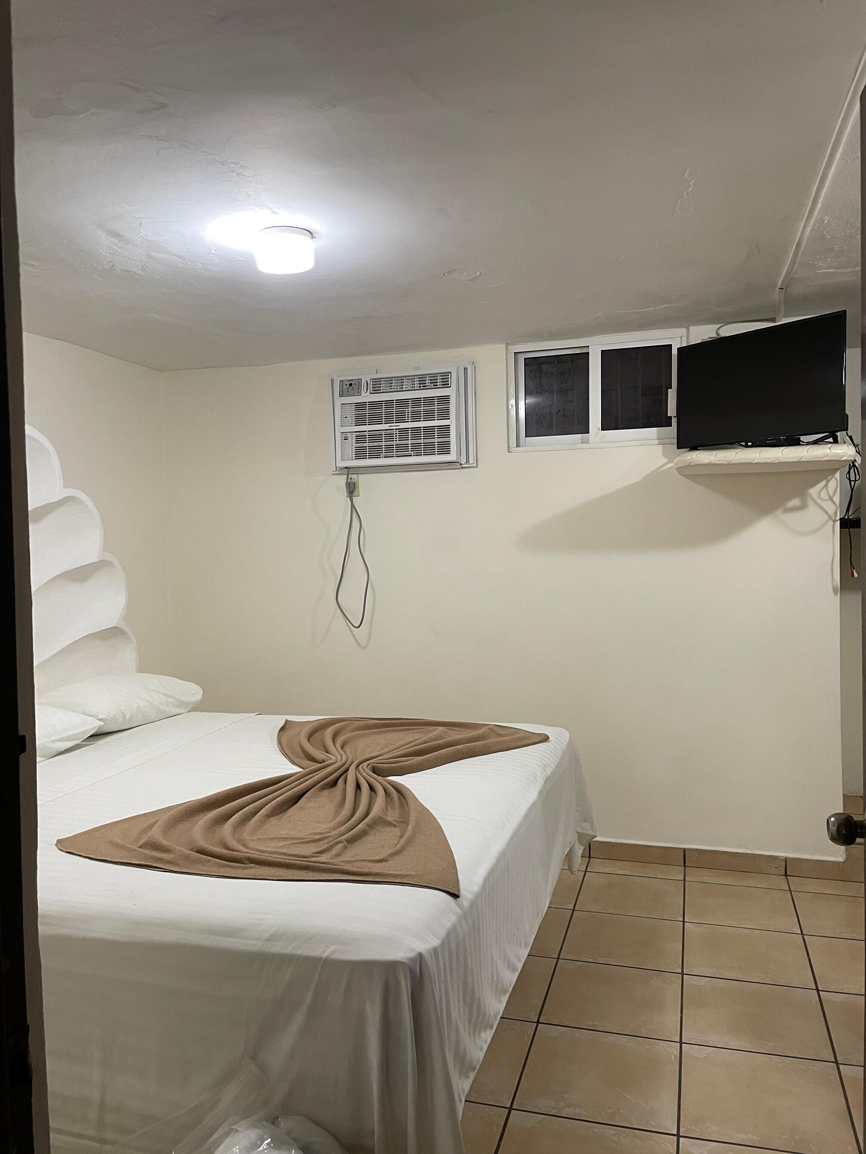 A bedroom with a bed, a TV, an air conditioning unit, and a window.
