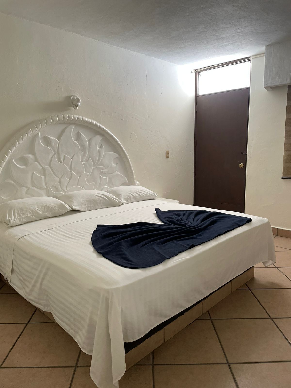 Un letto con una testiera bianca e una coperta blu scuro, una porta marrone e un pavimento piastrellato.