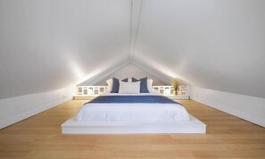 Ein Schlafzimmer mit schrägen Wänden, einem in der Mitte des Raumes stehenden Bett und einem weißen Geländer entlang des Deckenrandes.