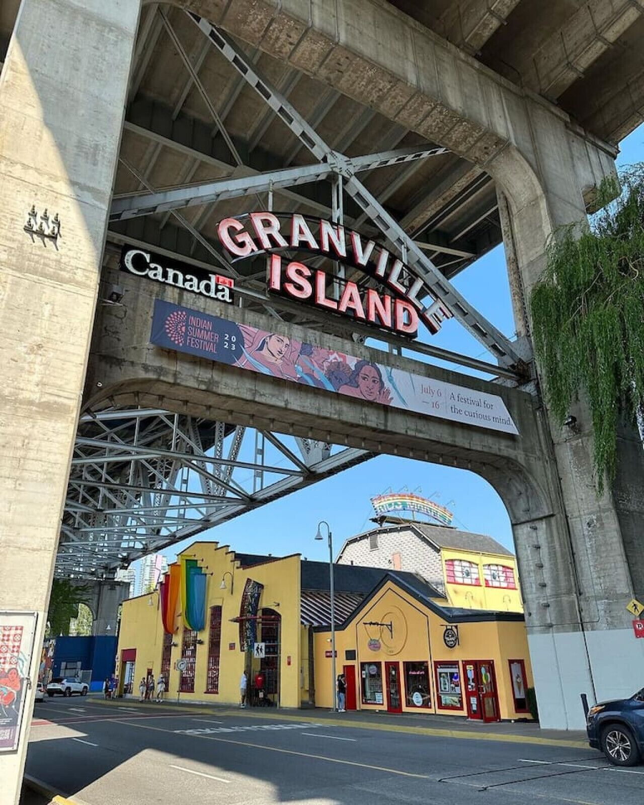 A large archway with 'Granville Island' signage, a yellow building with a red door, and a blue awning.