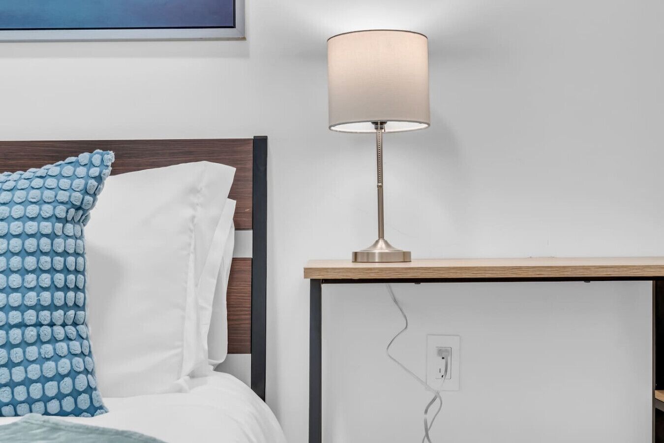 Un letto rifatto con cura, con un cuscino blu fantasia, uno bianco e una testiera in legno. Una moderna lampada da tavolo con paralume beige è posizionata su un comodino in legno accanto al letto.