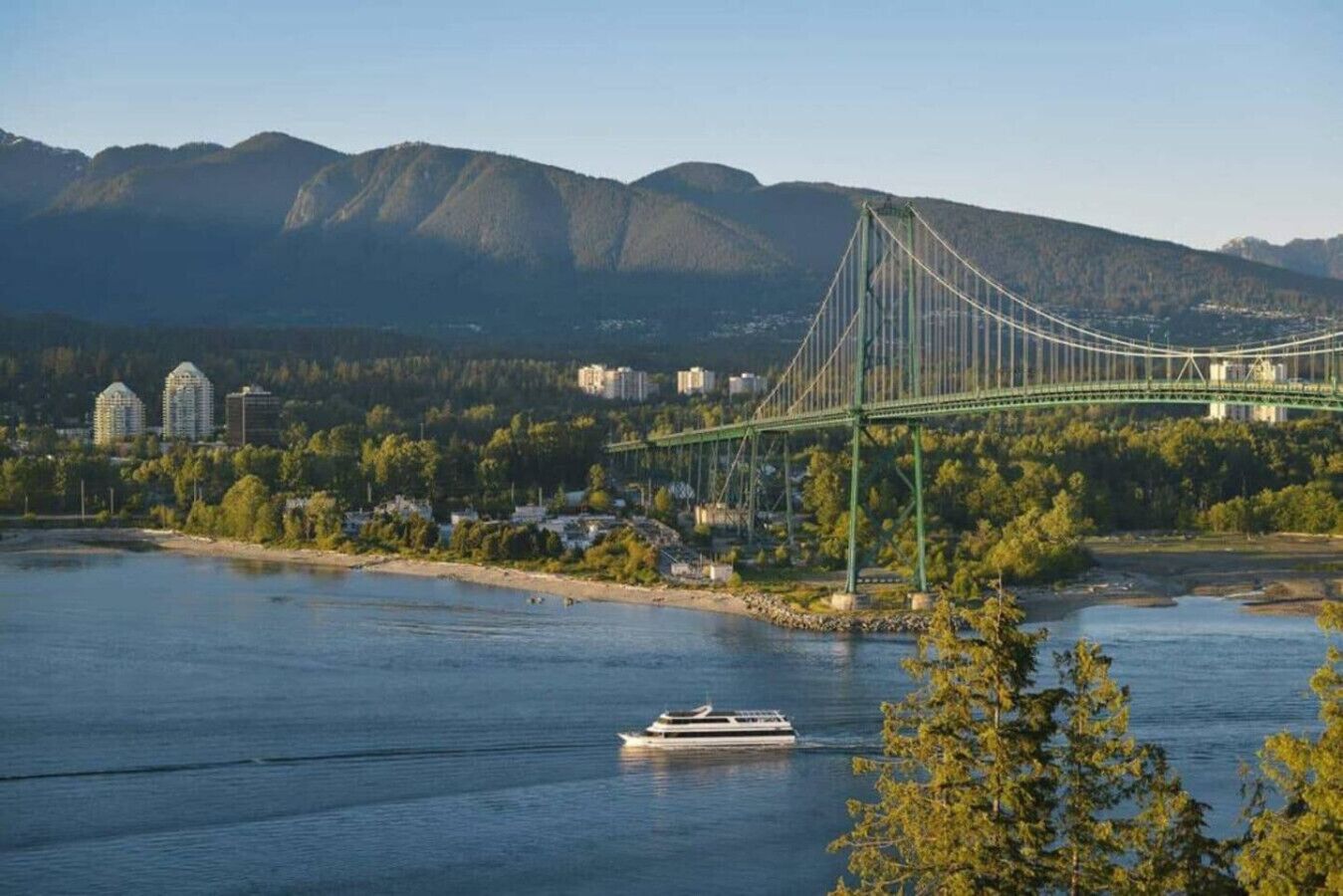 Een hangbrug over een rivier, met een boot, gebouwen en bergen op de achtergrond.