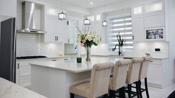 Una cocina moderna con armarios blancos, una campana extractora de acero inoxidable, luces colgantes, una isla central con taburetes y un televisor montado sobre el fregadero.