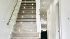Una escalera moderna con escalones de madera y barandillas de vidrio, que conduce a un pasillo bien iluminado con paredes y puertas blancas.
