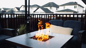 Una mesa cuadrada al aire libre con una chimenea en el centro, rodeada de mobiliario de mimbre y vistas a casas en la distancia.