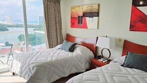 A hotel room with two beds, a ceiling fan, an air conditioning unit, a large window with a view of the city, and a painting on the wall. - Beach & Nightlife Paradise; Top floor and ocean view (Cancún)