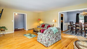 A living room with a floral sofa, wooden flooring, a dining area with chairs, and a chandelier. - 9 Mi to Lake George Village: Home in Queensbury (Queensbury)
