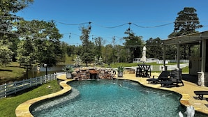 A swimming pool with a waterfall feature, surrounded by outdoor seating and a white fence. - Charming Farmhouse, Hot Tub, Pool, Outdoor Kitchen, Farm animals & Stocked Pond. (Diana)