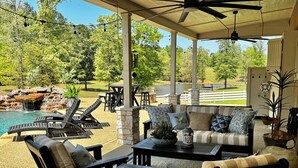An outdoor patio area with lounge chairs, a sofa, and a dining table. There is a pool with a waterfall feature in the background. - Charming Farmhouse, Hot Tub, Pool, Outdoor Kitchen, Farm animals & Stocked Pond. (Diana)