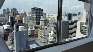 Une vue sur un paysage urbain à travers une fenêtre, avec de nombreux bâtiments, dont un gratte-ciel imposant et plusieurs immeubles de taille moyenne.