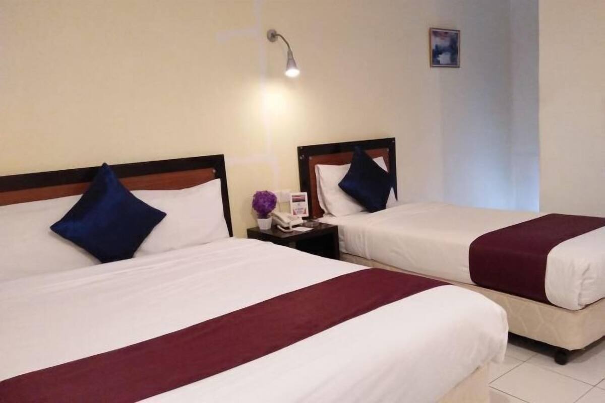Ein Hotelzimmer mit zwei Betten, jeweils mit einem hölzernen Kopfteil, weißer Bettwäsche und einem roten Akzent. Ein Nachttisch mit Lampe und einer kleinen Blumenvase steht zwischen den Betten.