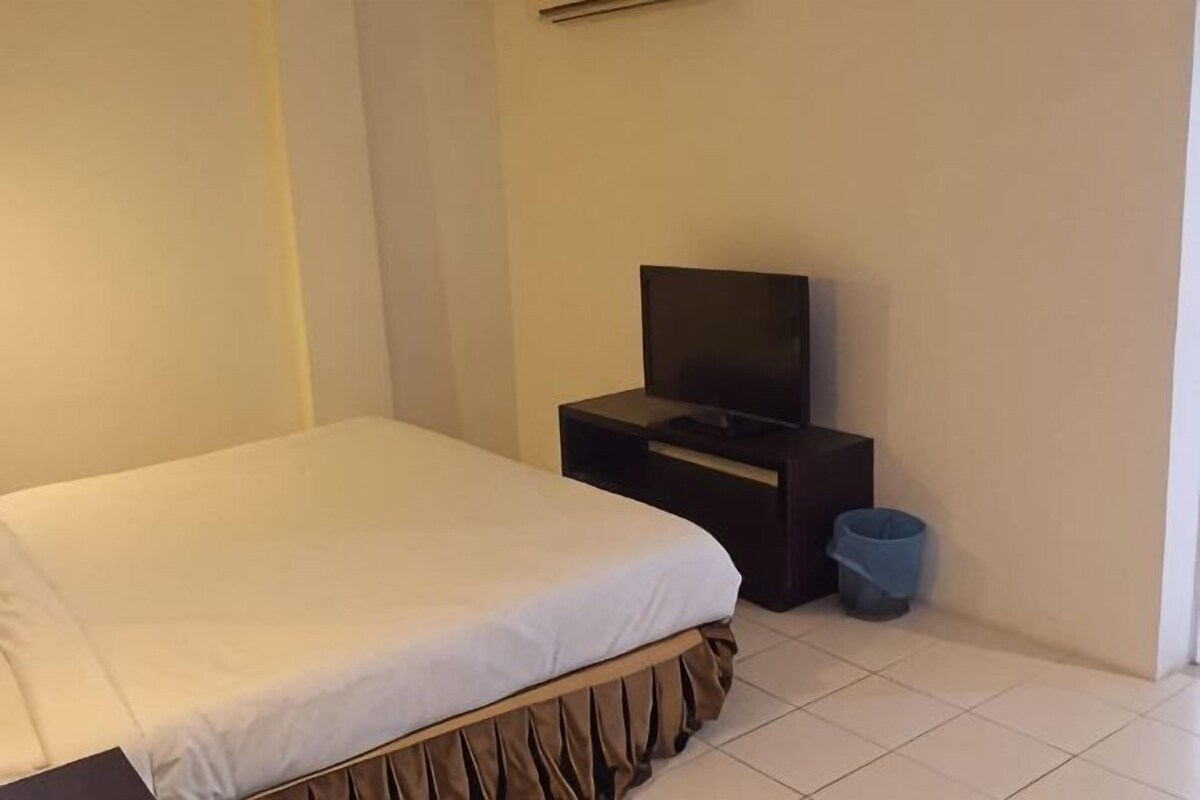 Habitación de hotel con cama, televisor sobre un mueble, un cubo de basura pequeño y piso de baldosas.