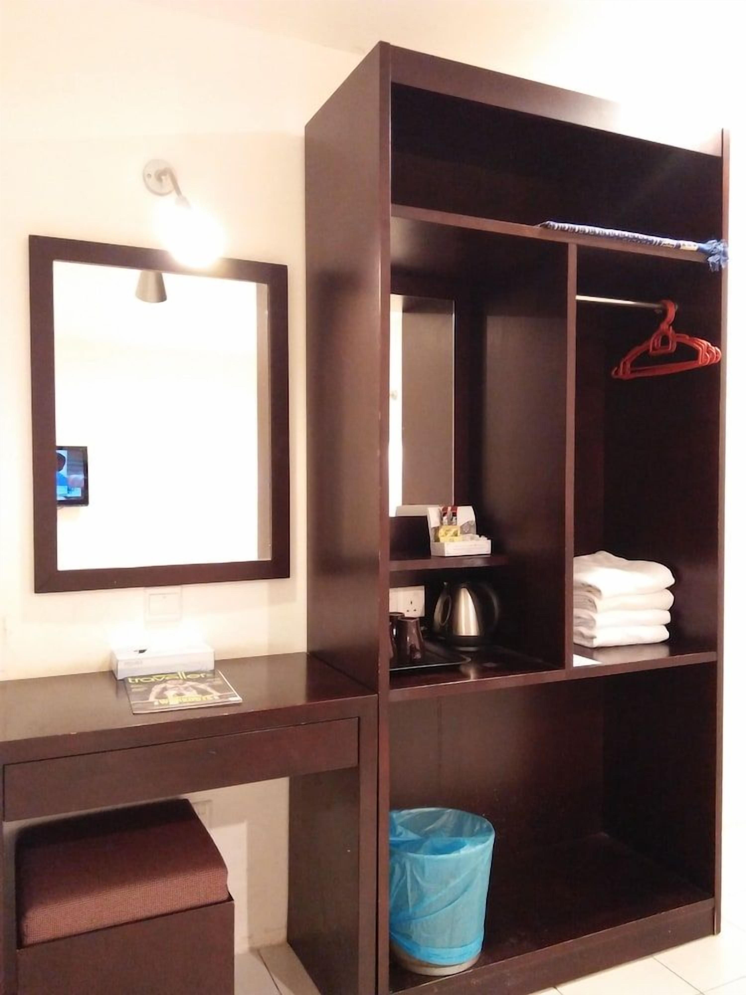 A room with a large wardrobe, a desk with a lamp, a mirror, and a trash bin.