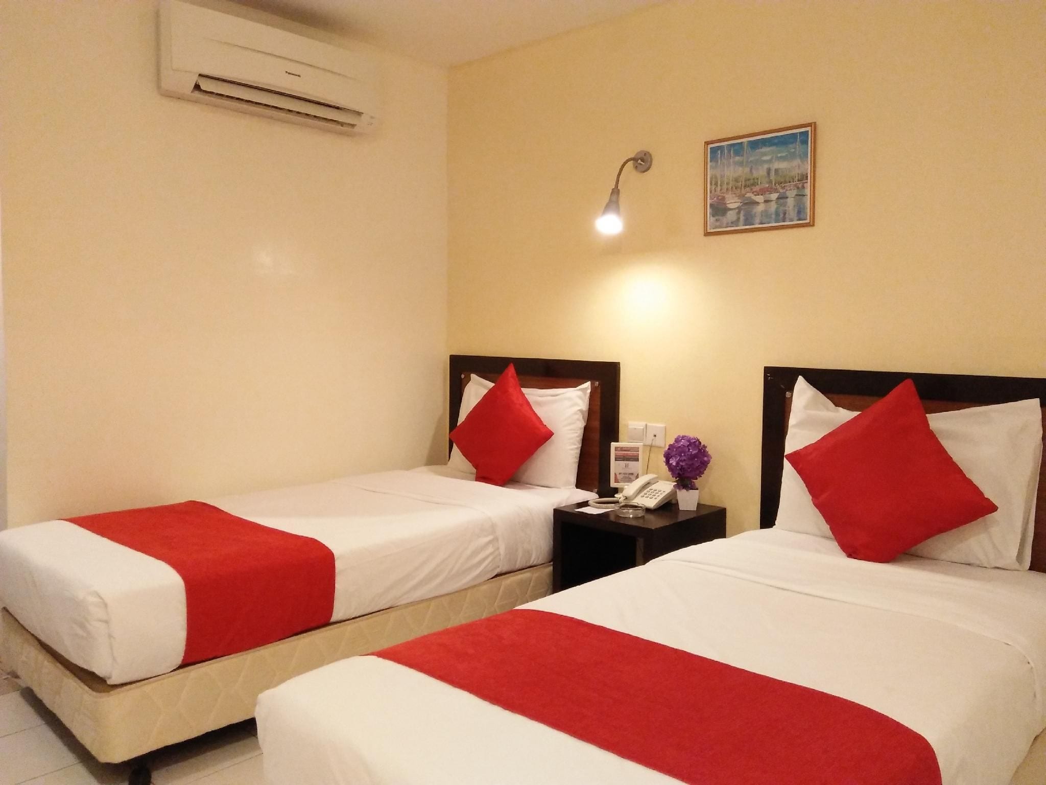 Une chambre d’hôtel avec deux lits, chacun recouvert d’une courtepointe rouge et blanche, une table de chevet avec un téléphone et un vase de fleurs, et un appareil de climatisation fixé au mur.