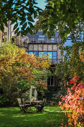 A garden with a wooden table and chairs, surrounded by lush greenery and autumn foliage. - The Hide (Ghent)
