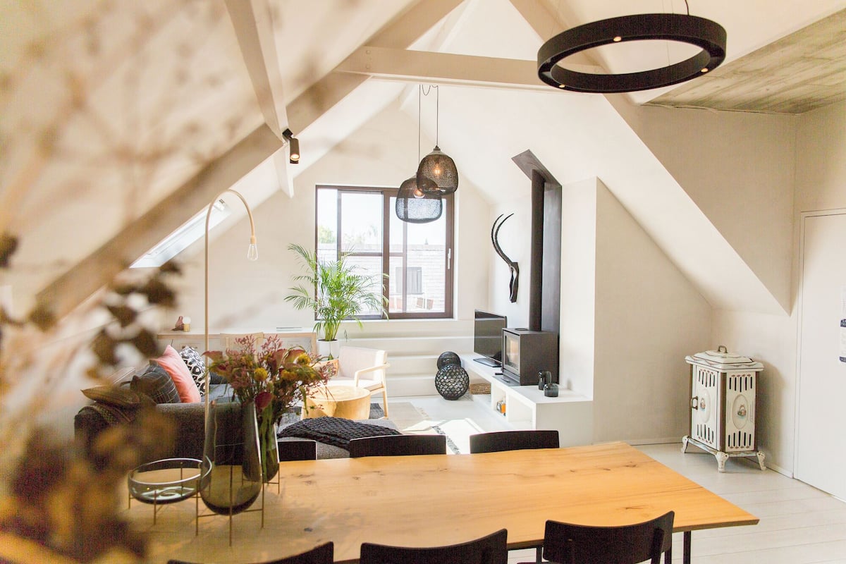 A modern loft with a high ceiling, exposed beams, a wooden dining table, and a large window providing natural light.