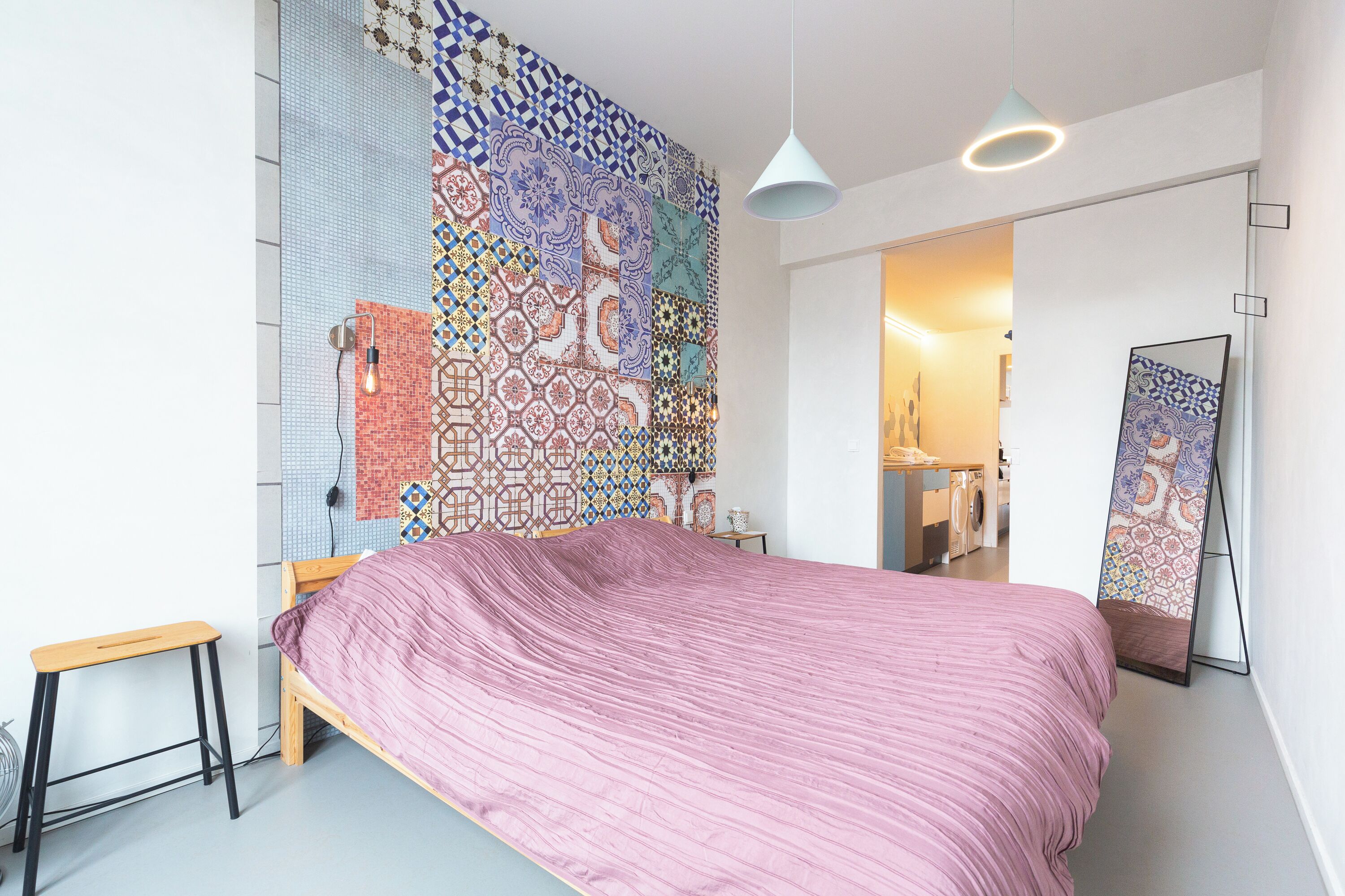 A bedroom with a large bed, a colorful mosaic wall, a small stool, and a hanging light fixture.