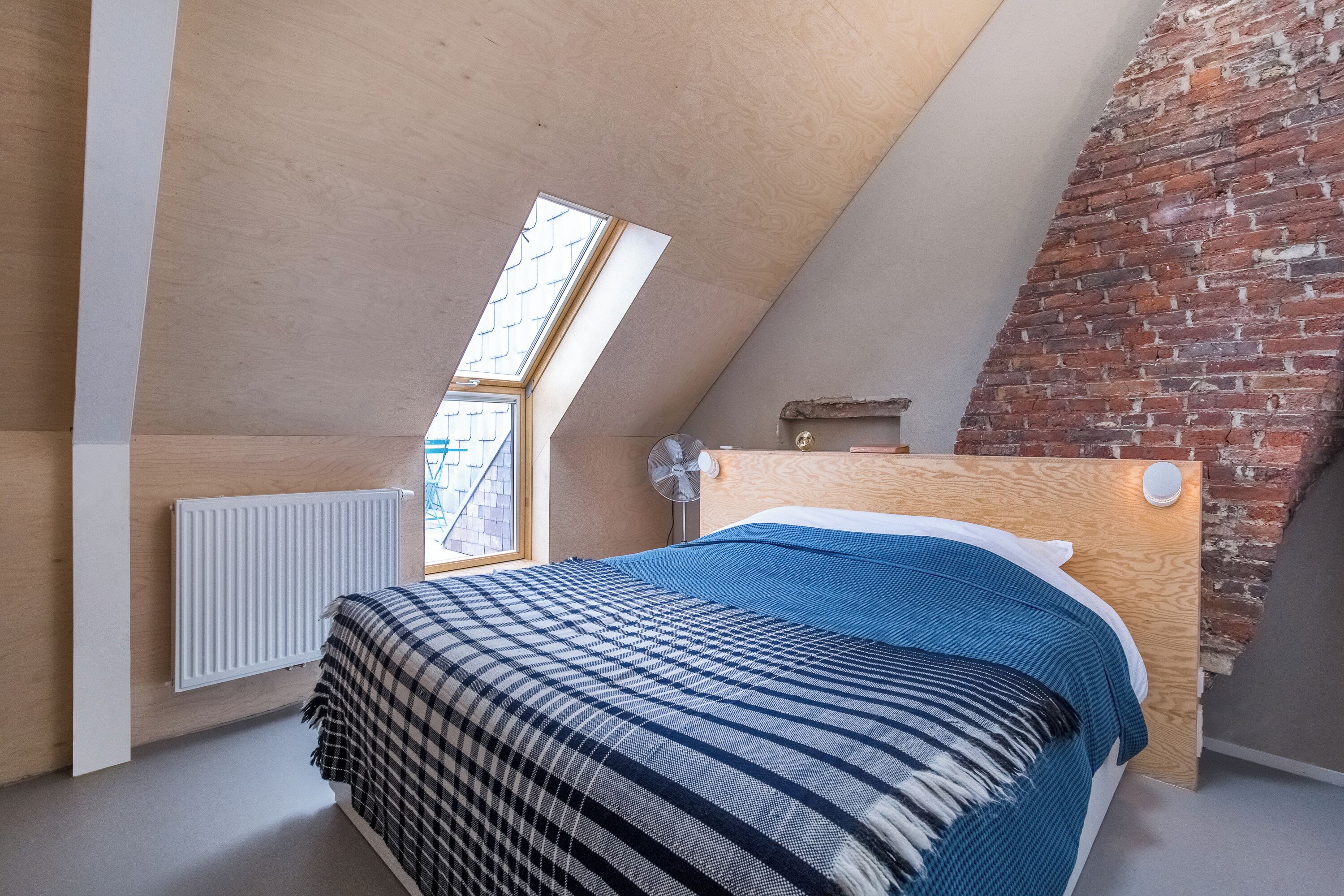 Een slaapkamer met een schuine plafond, een houten hoofdbord, een raam, een radiator, en een bed met een gestreept dekbed.