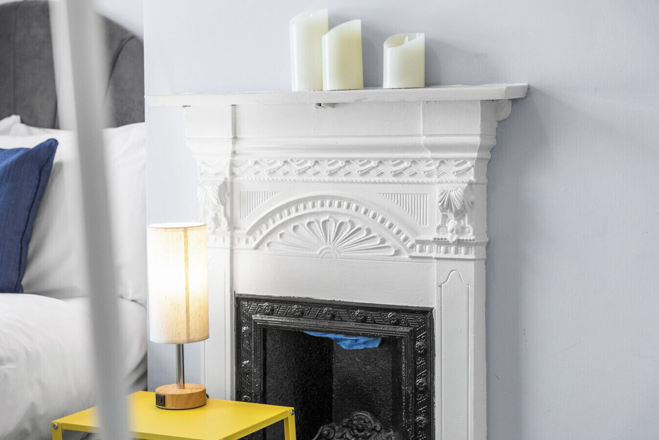 A white fireplace with three candles, a blue pillow, and a yellow nightstand with a lamp.