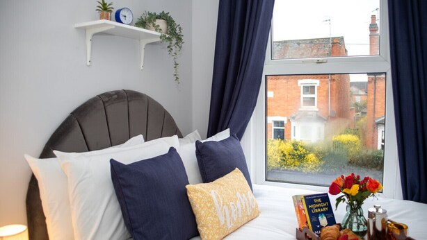 A neatly made bed with pillows, a bedside table with a book and a tray of food, a window with curtains, and a wall-mounted shelf with plants and a clock. - Charming 5-Bed Victorian House (Worcestershire)
