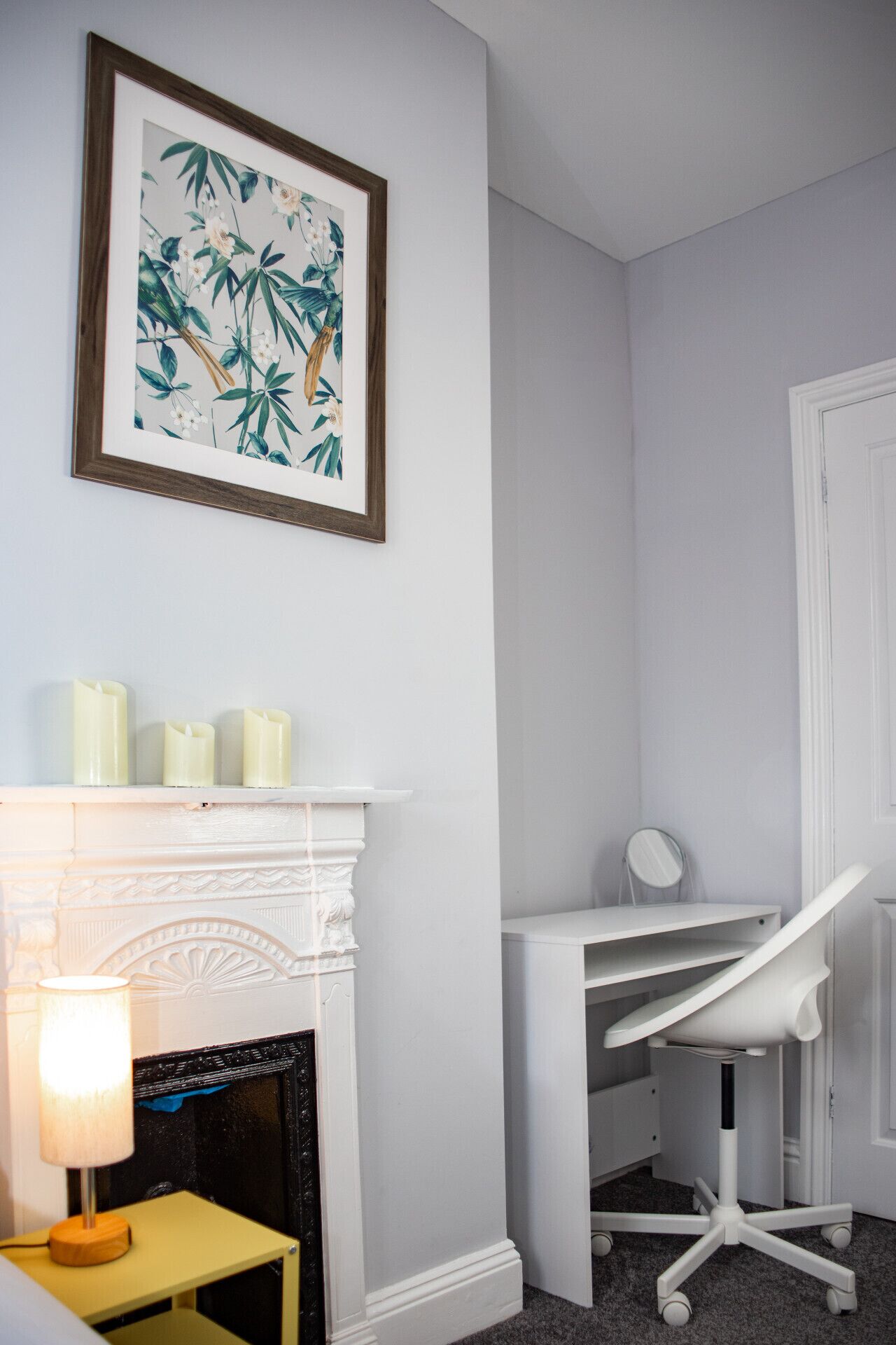 Een kamer met een witte open haard, een bureau met een stoel, en een lijst met een botanische afbeelding aan de muur.