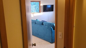 Un pasillo que conduce a una sala de estar con un sofá azul, un televisor montado en la pared y una ventana con cortinas estampadas.