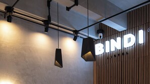 Un comptoir d’accueil moderne avec une enseigne indiquant « BINDI HOSTEL », deux tabourets blancs et un éclairage suspendu.