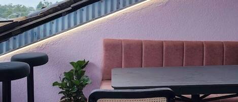 Una terraza en la azotea con una mesa, sillas, una planta en maceta y un banco con cojín rosa.