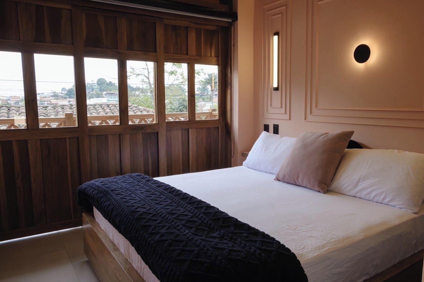 A bedroom with a large bed, wooden paneling, a view through the window, and a wall-mounted light fixture.