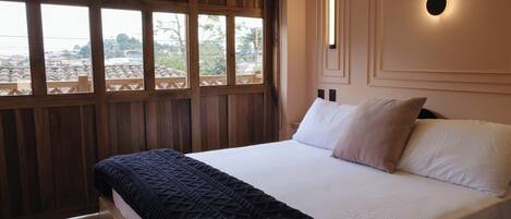 A bedroom with a large bed, wooden paneling, a view through the window, and a wall-mounted light fixture.
