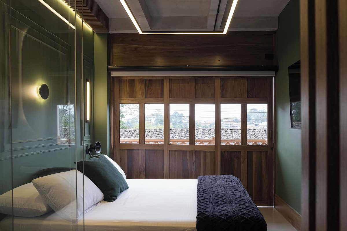A modern bedroom with a large bed, a glass partition, and a view of a cityscape through wooden panels.