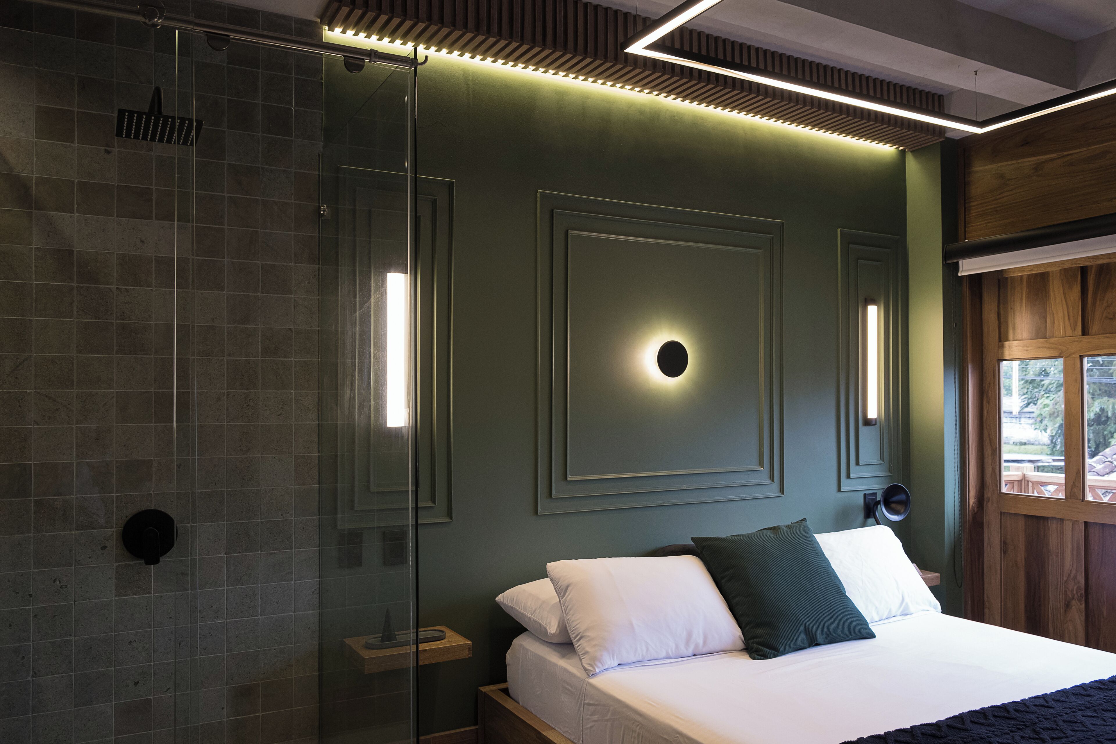 A modern bedroom with a glass shower enclosure, a bed with white and dark green bedding, and a wooden headboard.