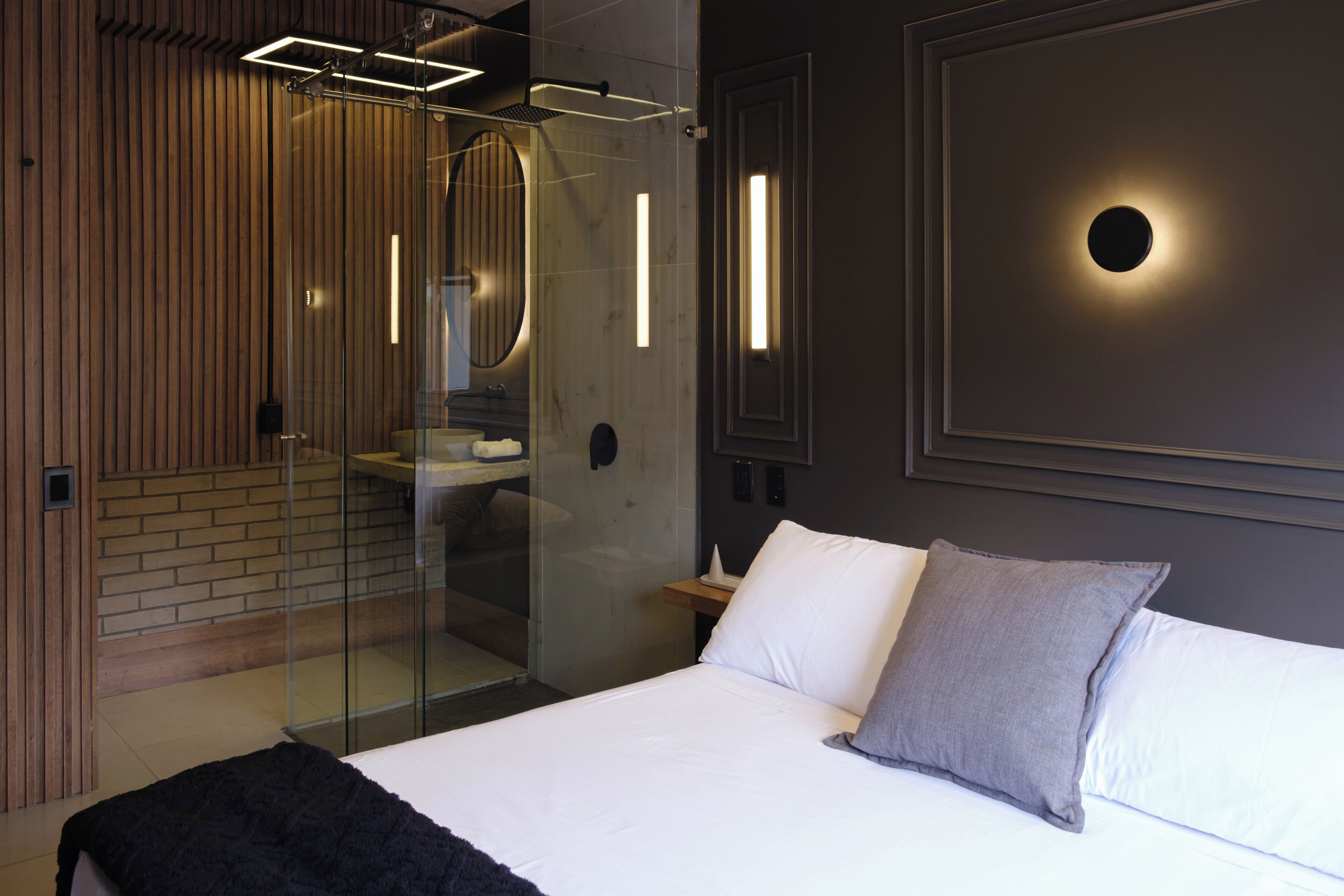 Une chambre moderne dotée d’une cabine de douche en verre, d’un grand lit avec une literie blanche et grise, et d’un luminaire fixé au mur.