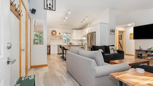 A modern living space with a grey sofa, wooden coffee table, and a kitchen area with white cabinets and a stainless steel refrigerator.