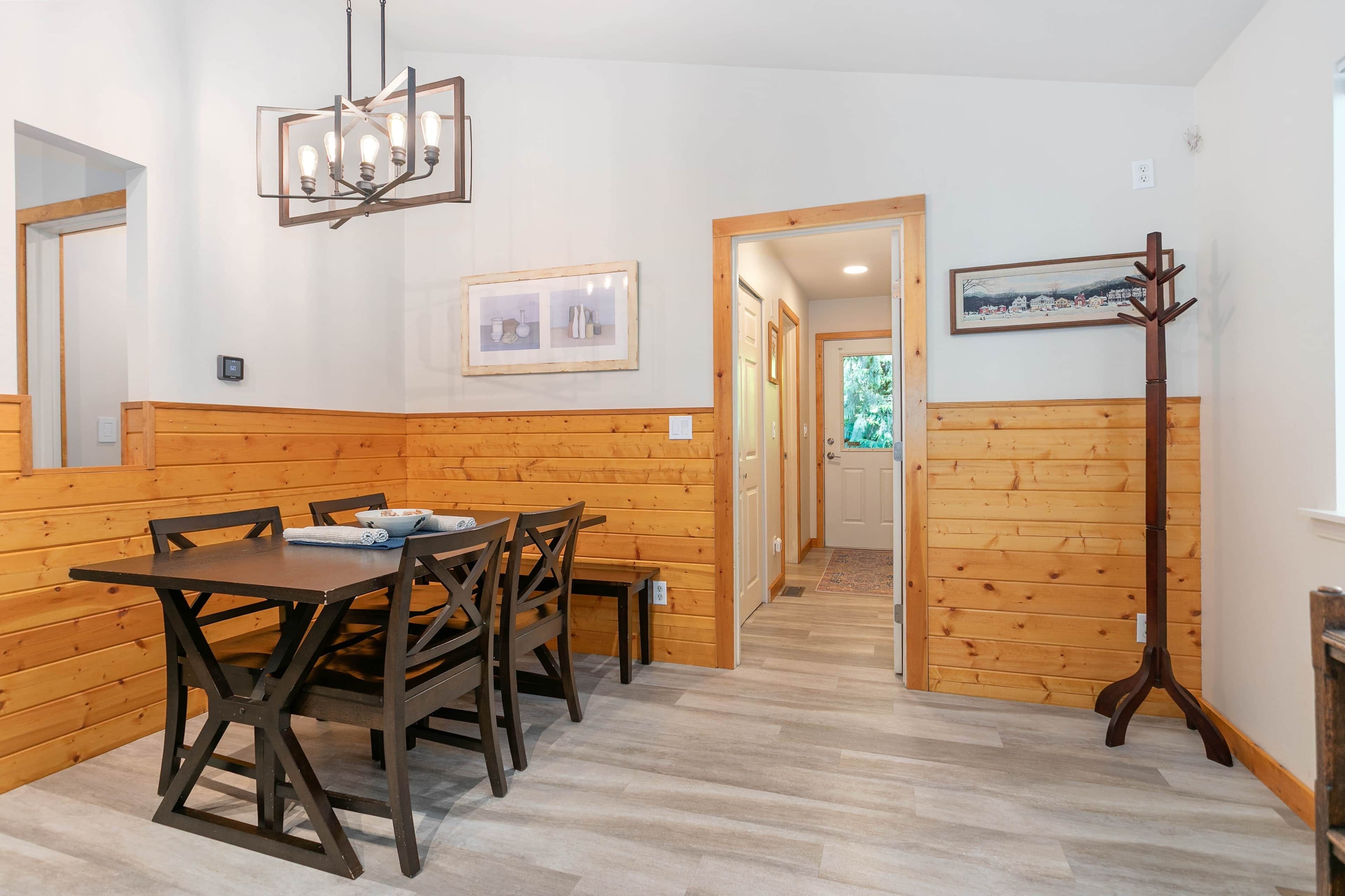 Une pièce aux murs lambrissés, comprenant une table à manger, des chaises, un banc, un porte-manteau et un couloir menant à une autre pièce.