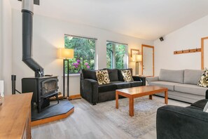 A living room with a wood-burning stove, two sofas, a coffee table, and large windows with a view of greenery. - Mt. Baker Cabin | Hot Tub | Fiber Internet | Generator (Deming)