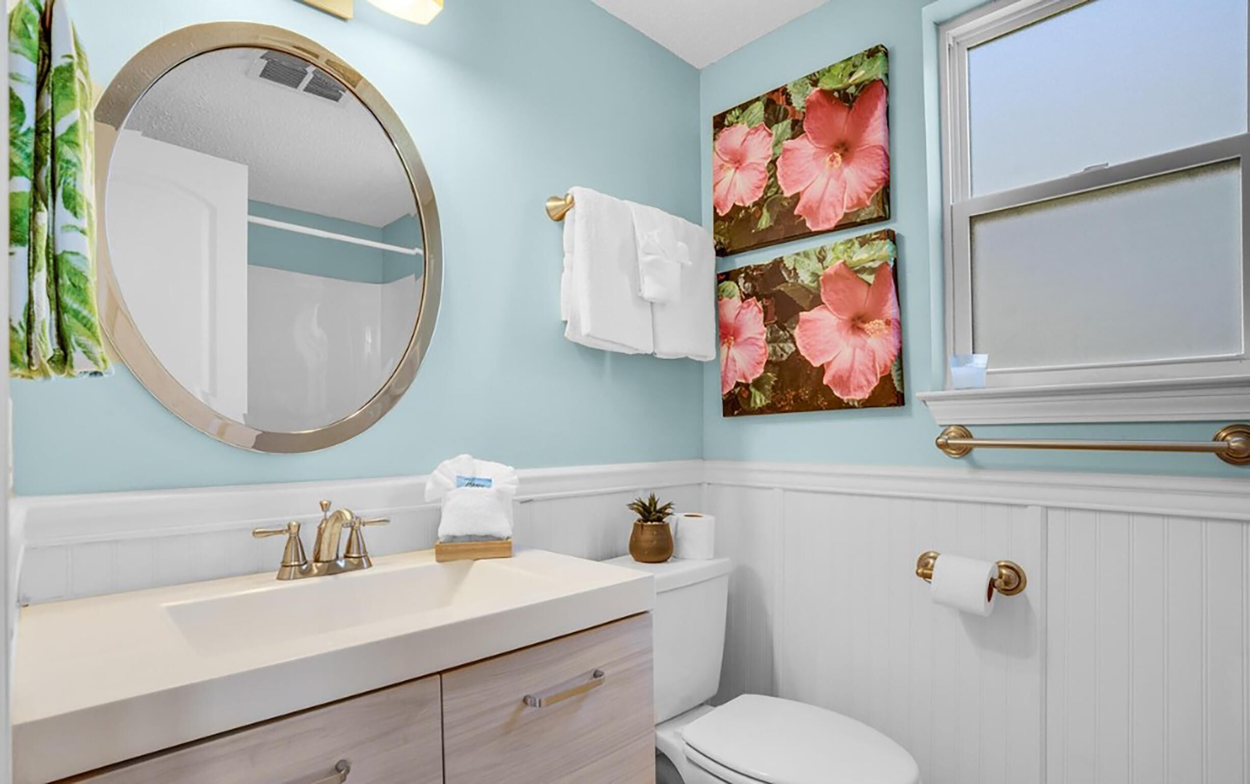 Un baño con un espejo redondo, un lavamanos con grifo dorado, un inodoro y dos obras de arte floral en las paredes.
