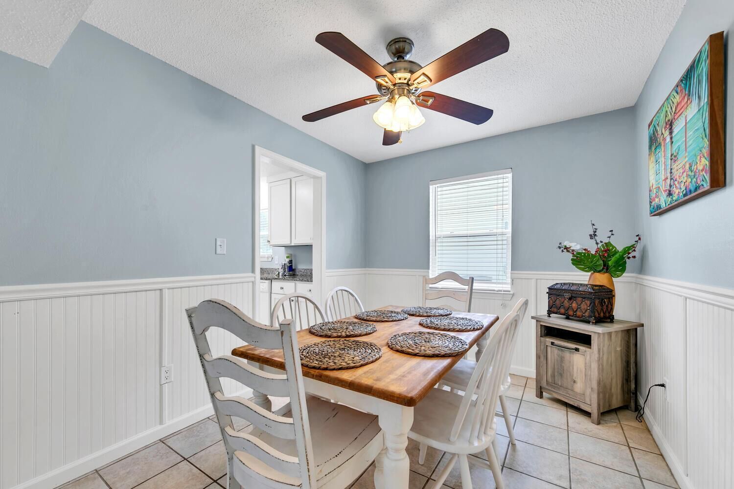 A dining area with a wooden table, white chairs, a ceiling fan, and a window with blinds.
