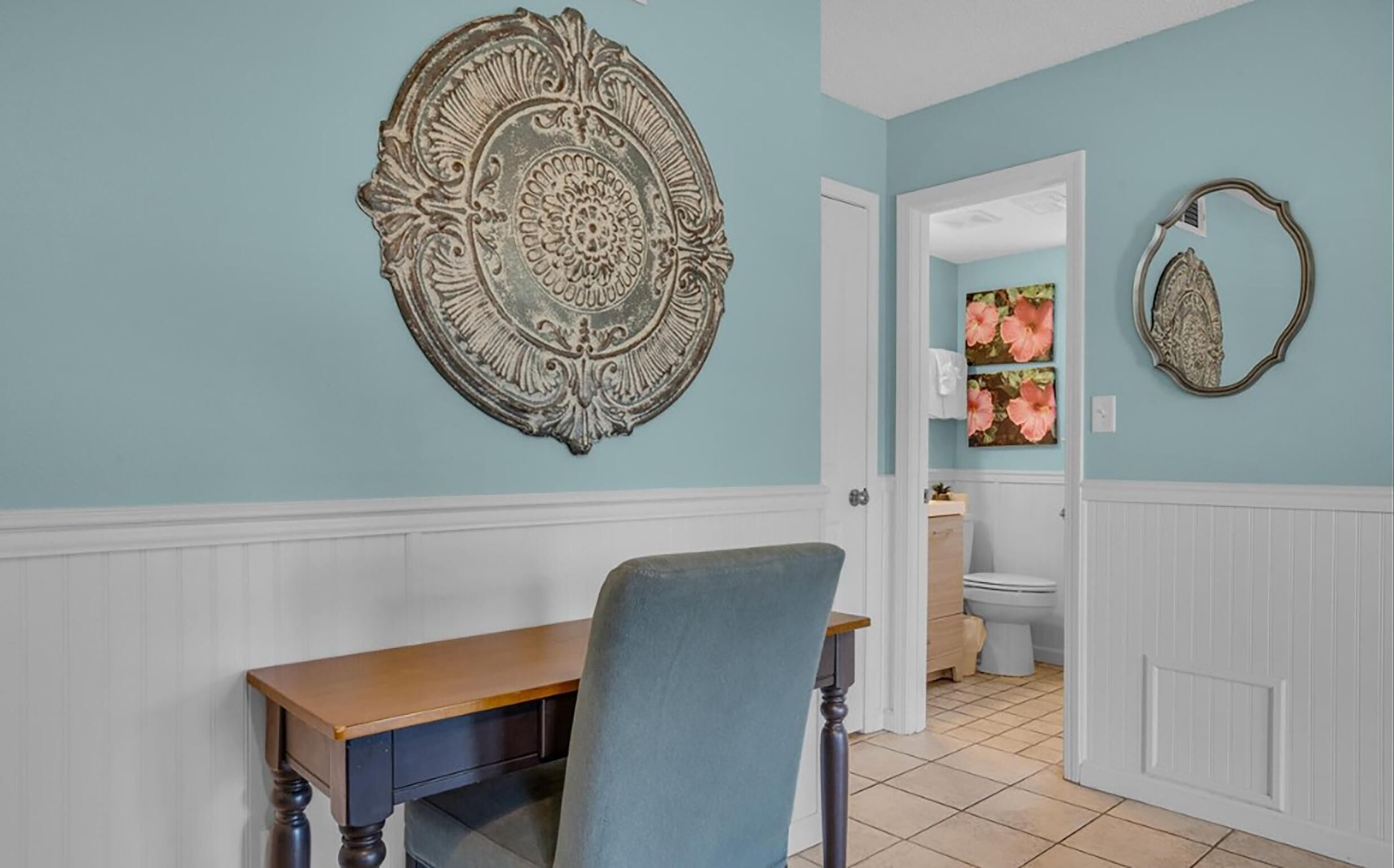 A room with a wooden table and a blue chair, a decorative wall piece, and a bathroom with a toilet and a sink.