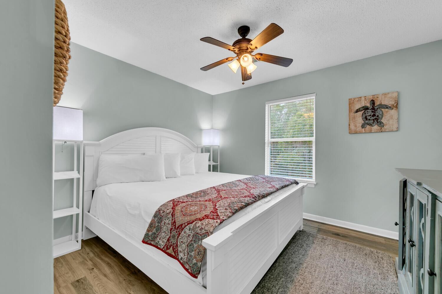 Une chambre avec un lit blanc, un ventilateur de plafond, une fenêtre avec des stores et une œuvre d’art accrochée au mur.