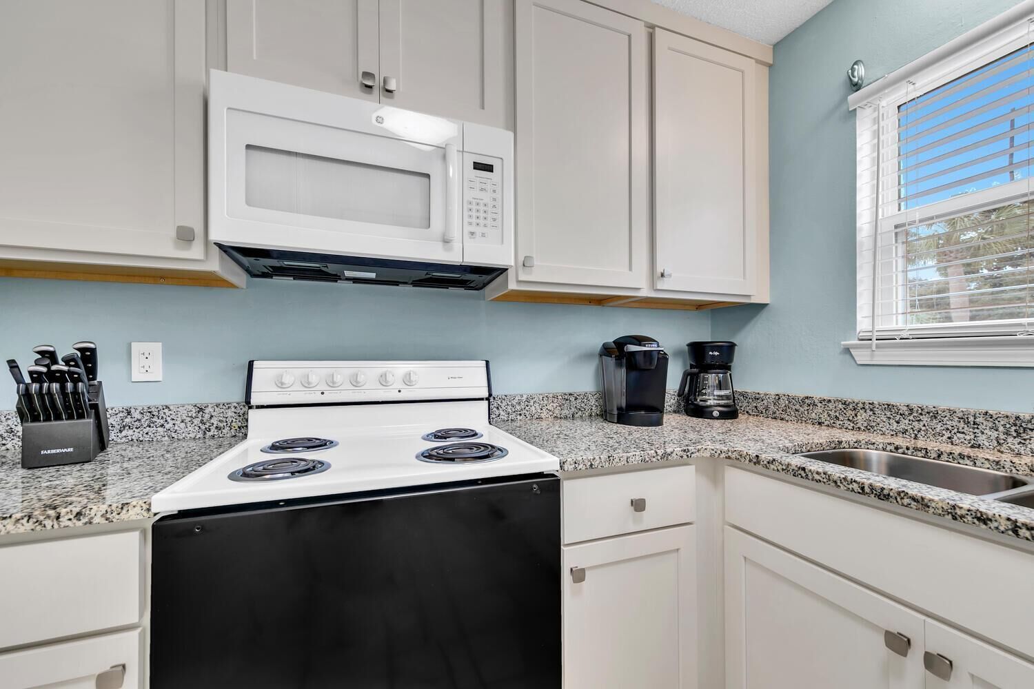 A kitchen with white cabinets, a white stove, a microwave, a coffee maker, and a sink.
