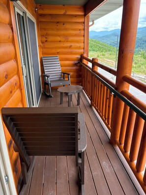 A wooden deck with a bench and two chairs, a table, and a railing overlooking a mountainous landscape.