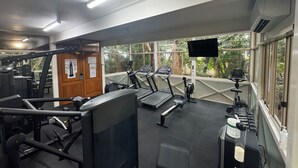 A gym with exercise equipment, a television, and large windows with a view of trees. - Apart King & Double Sofa Bed Kitchenette Heated Pool WIFI Big TV Walk to Beach (Port Douglas)